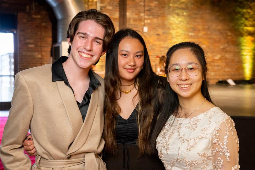 A young man wearing a beige coat, a young woman wearing a black shirt and a young woman wearing a white shirt smile